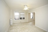 This single-floor space presents a clean, minimalist interior characterized by its all-white color palette and soft, neutral carpeting throughout. The main room is well-proportioned, offering a comfortable sense of openness while maintaining an intimate feel. Crown molding runs along the upper perimeter of the walls, adding a subtle architectural detail that elevates the overall aesthetic.

A ceiling fan with an integrated light fixture is centrally mounted, providing both air circulation and ambient lighting. Two windows positioned in the corner of the room allow natural light to filter in, brightening the space and creating an airy atmosphere. The windows are equipped with roller shades for privacy and light control, and a window-mounted air conditioning unit ensures year-round climate comfort.

A traditional white radiator sits beneath the windows, serving as the primary heat source and complementing the room's classic detailing. The smooth white walls and light carpeting work together to create a neutral, versatile backdrop well-suited for a variety of uses. Overall, the space conveys a tidy, functional character with thoughtful classic touches.