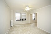 This single-floor space presents a clean, minimalist interior characterized by its all-white color palette and soft, neutral carpeting throughout. The main room is well-proportioned, offering a comfortable sense of openness while maintaining an intimate feel. Crown molding runs along the upper perimeter of the walls, adding a subtle architectural detail that elevates the overall aesthetic.

A ceiling fan with an integrated light fixture is centrally mounted, providing both air circulation and ambient lighting. Two windows positioned in the corner of the room allow natural light to filter in, brightening the space and creating an airy atmosphere. The windows are equipped with roller shades for privacy and light control, and a window-mounted air conditioning unit ensures year-round climate comfort.

A traditional white radiator sits beneath the windows, serving as the primary heat source and complementing the room's classic detailing. The smooth white walls and light carpeting work together to create a neutral, versatile backdrop well-suited for a variety of uses. Overall, the space conveys a tidy, functional character with thoughtful classic touches.