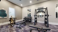 This fitness center offers a thoughtfully designed, single-floor layout that accommodates a comprehensive range of workout options. The space is finished with neutral, warm-gray walls and accented by a distinctive patterned carpet in muted purple and gray tones, lending a modern and polished aesthetic throughout.

The cardio zone features a row of treadmills, ellipticals, and a stair climber, each equipped with integrated digital display screens. Natural light filters in through multiple windows along the walls, creating a bright and energizing atmosphere, while framed artwork adds a tasteful decorative touch.

The strength training area is well-appointed with a Smith machine, a functional trainer cable system, a full dumbbell rack spanning a wide weight range, adjustable benches, and a multi-use functional training rig complete with TRX suspension straps and a variety of kettlebells and medicine balls. Wall-mounted mirrors in the free weights section allow users to monitor their form during exercises.

Additional amenities include a wall-mounted flat-screen television, recessed ceiling lighting, and a rowing machine for full-body conditioning. The space is accessible through glass-paneled doors and is equipped with ventilation to maintain a comfortable environment. Overall, this fitness center delivers a clean, modern, and fully functional setting suited for diverse training needs.