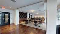 This single-floor workspace is thoughtfully designed to balance functionality with a refined aesthetic. Rich, dark hardwood flooring runs throughout the space, grounding the open layout and adding warmth to the overall atmosphere. The interior is divided into distinct yet connected work zones, fostering both individual focus and collaborative interaction.

The primary work area features multiple sleek, dark-topped tables paired with ergonomic rolling chairs in warm brown and mustard tones, arranged atop a neutral woven area rug that defines the space. A striking built-in bookcase with dark wood shelving lines one wall, offering ample storage and display space while contributing to the room's sophisticated character.

Toward the rear, a longer conference-style table is flanked by high-backed seating, providing a dedicated setting for meetings or group discussions. Pendant lighting and recessed ceiling fixtures cast a warm, layered glow throughout, complemented by decorative cage-style pendants that add a contemporary touch.

The white walls and structural columns contrast cleanly against the dark wood tones, creating a polished, professional environment that feels both welcoming and well-appointed.