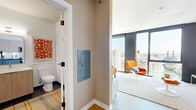Located in Chicago's West Loop neighborhood at 234 W Polk Street, this single-floor unit showcases a thoughtfully designed open layout with a bold, contemporary aesthetic. Exposed concrete ceilings lend an industrial character throughout, complemented by light wood-toned flooring that runs seamlessly from entry to living area.

The living space is defined by dramatic floor-to-ceiling windows that frame expansive views of the Chicago River and city skyline, flooding the interior with natural light. Vibrant orange accent walls and colorful furnishings create a dynamic, energetic atmosphere. A large-format cityscape artwork anchors the main living wall, reinforcing the urban setting.

The kitchen features a clean, modern design with two-tone cabinetry — light wood uppers paired with muted blue-gray lowers — dark countertops, a tile backsplash, and stainless steel appliances including a gas range and over-range microwave. A louvered closet door near the entry provides convenient storage access.

The bathroom is finished with a warm wood vanity, white quartz countertop, undermount sink, and a large frameless mirror, all set against crisp white walls. Colorful geometric artwork adds a distinctive decorative touch. The entry corridor connects all spaces efficiently, making the overall layout highly functional for urban living.
