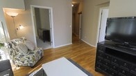 This single-story residence showcases a cohesive, functional layout centered around warm hardwood floors that extend throughout the main living areas and hallway, lending a sense of continuity and warmth to the space. The primary living area is comfortably sized, featuring neutral taupe-toned walls that create a calm, versatile backdrop suitable for a variety of interior styles. A central hallway connects the living space to additional rooms, offering an efficient and practical flow throughout the home.

Visible through one doorway is a carpeted room, likely a bedroom, which contrasts subtly with the hardwood in the main areas and provides a softer underfoot feel. Dark-finished furniture pieces, including multi-drawer dressers, are present in both the main room and the adjoining bedroom, adding a bold contrast against the lighter walls and flooring. Recessed ceiling lighting provides focused illumination in the hallway, while the overall lighting scheme throughout remains clean and understated.

White trim and door casings frame each entryway, adding a crisp, finished detail that complements the neutral palette. The space presents a straightforward, move-in-ready character with a practical single-floor design.
