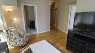 This single-story residence showcases a cohesive, functional layout centered around warm hardwood floors that extend throughout the main living areas and hallway, lending a sense of continuity and warmth to the space. The primary living area is comfortably sized, featuring neutral taupe-toned walls that create a calm, versatile backdrop suitable for a variety of interior styles. A central hallway connects the living space to additional rooms, offering an efficient and practical flow throughout the home.

Visible through one doorway is a carpeted room, likely a bedroom, which contrasts subtly with the hardwood in the main areas and provides a softer underfoot feel. Dark-finished furniture pieces, including multi-drawer dressers, are present in both the main room and the adjoining bedroom, adding a bold contrast against the lighter walls and flooring. Recessed ceiling lighting provides focused illumination in the hallway, while the overall lighting scheme throughout remains clean and understated.

White trim and door casings frame each entryway, adding a crisp, finished detail that complements the neutral palette. The space presents a straightforward, move-in-ready character with a practical single-floor design.