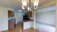 This single-floor residence offers a practical and cohesive layout centered around an open-concept kitchen and living area. The kitchen is equipped with medium-toned wood cabinetry, dark granite-style countertops, and black appliances, including a refrigerator and range. A peninsula serves as a natural divider between the kitchen and the adjacent living space, providing additional counter space and a convenient casual dining or prep area. Hardwood-style flooring runs throughout the kitchen and entry zones, transitioning to plush carpeting in the living area, adding a layer of warmth and comfort underfoot. Overhead, a multi-arm chandelier with frosted glass shades anchors the dining area, providing a decorative focal point while offering ample illumination. A flush-mount ceiling light near the entryway ensures the space is well-lit throughout. The textured ceiling adds subtle depth to the interior. The front entry door is clearly visible from the kitchen, contributing to an open and connected floor plan that promotes easy movement between functional zones. Overall, the space presents a straightforward, functional design with classic finishes throughout.
