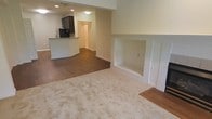This single-floor space showcases an open-concept layout that seamlessly connects the living area to the kitchen. The living room features plush, neutral-toned carpeting that transitions into hardwood-style flooring, creating a natural visual distinction between living and dining or kitchen zones. A standout architectural detail is the built-in fireplace niche, framed by clean white millwork with fluted column accents, adding a classic focal point to the room.

The kitchen is positioned toward the rear of the open floor plan and is equipped with dark cabinetry, a peninsula-style breakfast bar with dark countertops, and recessed lighting overhead. The bar provides a casual dining or prep area while maintaining the open sightlines throughout the space. A doorway visible beyond the kitchen suggests access to additional rooms or a pantry area.

The walls are finished in a soft, neutral beige tone throughout, contributing to a bright and cohesive ambiance. Recessed lighting in the kitchen and flush-mount fixtures provide consistent illumination. The overall design balances functional living with understated, classic detailing.