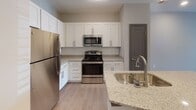 This single-floor residence at 101 Three Mile Loop in Kannapolis, North Carolina offers a thoughtfully designed layout with modern finishes throughout. The kitchen serves as a standout feature, equipped with stainless steel appliances including a refrigerator, range, and microwave, complemented by crisp white shaker cabinetry, granite countertops with a subtle speckled pattern, a subway tile backsplash, and a large kitchen island with a double stainless steel sink and pendant lighting overhead. Light wood-style flooring runs through the kitchen and main living areas, adding warmth and continuity to the space. The living area is generously sized, featuring a ceiling fan, recessed lighting, and a sliding glass door that opens to an outdoor balcony, allowing natural light to fill the room. Bedrooms are finished with plush wall-to-wall carpeting and include windows with blinds that bring in additional natural light. The bathroom is well-appointed with granite countertops, a white vanity with brushed nickel hardware, a large mirror, and a tub-shower combination. Neutral gray tones on the walls throughout the unit create a clean, contemporary aesthetic, while dark baseboards provide a refined contrast to the light flooring and walls.