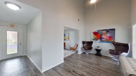This single-floor space presents a thoughtfully designed interior characterized by soaring vaulted ceilings, wide-plank hardwood-style flooring, and crisp white walls that create a bright, airy atmosphere throughout. The layout flows through several distinct open zones, each offering its own character while maintaining a cohesive contemporary aesthetic.

A welcoming entry corridor leads into the main areas, where the ceiling height dramatically increases, amplifying the sense of openness. One prominent gathering area features a bold navy blue accent wall rising to meet the vaulted ceiling, anchored by large windows that admit generous natural light and views of the outdoors. A ceiling fan adds functionality to this well-lit space.

Adjacent rooms offer additional flexible areas for seating or focused use, with partitioned wall openings creating visual connectivity between zones without sacrificing definition. A pass-through counter suggests a kitchen or service area nearby, fitted with modern fixtures.

Throughout, the space is accented with vibrant contemporary artwork, lending a creative and dynamic energy. The exit corridor, equipped with safety features, provides a well-lit secondary access point. Overall, the space combines architectural interest, natural light, and versatile open zones to form a stylish and functional environment.