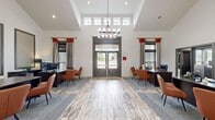 This single-story community clubhouse and leasing office offers a thoughtfully designed, multi-functional space spanning a generous footprint across five distinct areas. The grand reception lobby immediately impresses with soaring vaulted ceilings, an abundance of natural light streaming through a clerestory window bank above the main entrance, and a striking chandelier as a central focal point. Wood-look tile flooring runs throughout, complemented by patterned area rugs that define workstation zones where dark espresso desks and cognac leather chairs create a professional yet welcoming atmosphere. Bold red drapery accents add a vibrant pop of color against the crisp white walls. The adjacent lounge area features vaulted ceilings, comfortable seating, decorative wall art, and views to the outdoors, providing a relaxed gathering space. The fully equipped kitchen showcases dark cabinetry with granite countertops, stainless steel appliances, a mosaic tile backsplash, and a large island with bar seating — ideal for community events. A dedicated business center offers built-in granite-topped workstations, computer equipment, and a bold two-tone navy and white color scheme, providing a focused environment for residents. Together, these spaces combine modern design with practical amenities to create a vibrant community hub.