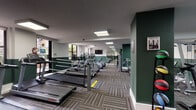 This fitness center is a comprehensively outfitted single-floor facility designed to support a wide range of workout routines. The space is defined by a distinctive two-tone color scheme, with deep forest green accent walls contrasting against crisp white upper walls, creating a vibrant and energizing atmosphere throughout.

The cardio zone is generously sized, housing a row of Life Fitness treadmills, elliptical trainers, and stair climbers aligned along large windows that invite ample natural light and offer views of the surrounding exterior. A wall-mounted flat-screen television provides entertainment for cardio sessions.

A dedicated cycling area features multiple Peloton-style stationary bikes arranged in front of a full-length mirror, which enhances the sense of space and allows users to monitor their form. Alongside the bikes, a medicine ball rack stocked with a variety of weighted balls adds functional training options to this zone.

The strength training section includes a multi-use cable and weight machine, as well as a rowing machine, catering to both resistance and full-body conditioning workouts. The floor throughout is covered in durable, patterned carpet tiles that provide both comfort and a polished aesthetic. Recessed and flush-panel ceiling lights ensure bright, consistent illumination across the entire facility, making it a practical and well-designed space for fitness.