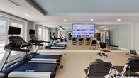 This fitness room in Naperville, IL, is a thoughtfully designed, single-floor facility offering a comprehensive workout environment. The space is finished with neutral carpeting, soft gray walls, and recessed lighting complemented by large ceiling fans, creating a comfortable and well-ventilated atmosphere.

A defining architectural feature is the expansive mirrored wall, which visually enlarges the space and enhances functionality for exercise form and technique. Mounted directly on the mirror is a large flat-screen television, providing access to on-demand workout content. French doors along an exterior wall invite natural light and offer outdoor views, further brightening the interior.

The cardio zone includes multiple treadmills and upright and recumbent stationary bikes, while the strength and functional training area is equipped with a cable machine, medicine ball rack, resistance bands, exercise mats, stability balls, and pilates-style chairs. A wall-mounted clock keeps users on schedule.

The layout flows naturally between cardio and functional training zones, maximizing usability. An adjacent room, visible through a doorway, suggests additional support facilities nearby. Overall, the space delivers a clean, organized, and fully appointed fitness environment suited for a wide range of exercise routines.