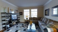 This two-floor home offers a well-appointed living space that blends comfort and style. The living room serves as a central gathering area, highlighted by two large windows that invite abundant natural light and offer views of the surrounding exterior. Crown molding along the ceiling adds an elegant architectural touch, while neutral wall tones create a warm, versatile backdrop. A large sectional sofa in a light gray upholstery anchors the seating area, complemented by a tan accent sofa adorned with decorative pillows in earthy tones. A glass-top coffee table sits atop a patchwork-style area rug that introduces a mix of textures and subtle patterns to the floor. An open shelving media unit along one wall provides display space alongside the mounted flat-screen television, while framed artwork adds character to the walls. Decorative ceramic vases in amber tones on the coffee table introduce a pop of color. The overall ambiance of the living area is inviting and tastefully curated, balancing functional furniture arrangements with decorative elements. The space extends across two floors, offering additional living and private areas throughout the home.