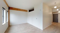 This single-storey space showcases a minimalist design characterized by crisp white walls and light-toned laminate or vinyl plank flooring that lends a clean, contemporary feel throughout. A standout architectural detail is the exposed natural wood beam running along the upper portion of the room, introducing a warm, organic contrast against the otherwise neutral palette. The ceiling height is notably generous, contributing to an open and airy atmosphere. A partial wall or structural divider is visible within the space, creating subtle separation and adding architectural interest without compromising the open feel. Near the top of this divider, a recessed opening adds a design element that enhances visual depth. The walls are fitted with standard electrical fixtures and a wall-mounted control panel, suggesting practical, move-in-ready functionality. White baseboards frame the flooring neatly, reinforcing the space's clean, well-finished appearance. Overall, the interior presents a bright, neutral canvas that is well-suited for a variety of uses, combining modern minimalism with the natural warmth of exposed wood accents.