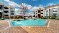 This outdoor pool area is located within a well-maintained residential community in Kenner, Louisiana. The centerpiece of the space is a generously sized swimming pool with clear, blue water, surrounded by a broad brick paver deck that provides ample room for lounging and socializing. The pool's geometric shape and clean tile border give it a classic, well-appointed appearance.

The surrounding grounds are thoughtfully landscaped with a mix of manicured shrubs, ornamental grasses, and mature trees that offer natural character and shade throughout the property. A dedicated grilling and outdoor cooking station is positioned along the perimeter, making the area well-suited for outdoor gatherings. Picnic-style seating is also available nearby, adding to the communal atmosphere.

The pool area is enclosed by a black iron fence, providing both security and an open, airy feel. A paved walkway lined with lush greenery connects the pool to the adjacent residential buildings, which feature a traditional architectural style with neutral siding, pitched rooflines, and multi-story balconies. Large windows on the surrounding structures allow for views of the outdoor amenity space. The open sky and abundant natural light further enhance the inviting, resort-like ambiance of this community outdoor space.