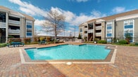 This outdoor pool area is located within a well-maintained residential community in Kenner, Louisiana. The centerpiece of the space is a generously sized swimming pool with clear, blue water, surrounded by a broad brick paver deck that provides ample room for lounging and socializing. The pool's geometric shape and clean tile border give it a classic, well-appointed appearance.

The surrounding grounds are thoughtfully landscaped with a mix of manicured shrubs, ornamental grasses, and mature trees that offer natural character and shade throughout the property. A dedicated grilling and outdoor cooking station is positioned along the perimeter, making the area well-suited for outdoor gatherings. Picnic-style seating is also available nearby, adding to the communal atmosphere.

The pool area is enclosed by a black iron fence, providing both security and an open, airy feel. A paved walkway lined with lush greenery connects the pool to the adjacent residential buildings, which feature a traditional architectural style with neutral siding, pitched rooflines, and multi-story balconies. Large windows on the surrounding structures allow for views of the outdoor amenity space. The open sky and abundant natural light further enhance the inviting, resort-like ambiance of this community outdoor space.