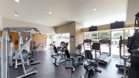 This fitness center is a thoughtfully designed, single-floor facility built to support a comprehensive workout experience. The space is outfitted with a diverse selection of cardio equipment, including treadmills, stationary bikes, and elliptical machines, all arranged to maximize floor space and ease of movement. A dedicated strength training area features a multi-function cable machine and free weight stations, catering to a full range of exercise routines. The interior design features warm, neutral-toned walls complemented by a light ceiling with recessed lighting, creating a bright and energizing atmosphere. Large windows line the perimeter, allowing abundant natural light to fill the room while offering views of a nearby outdoor pool and lush surrounding trees. The elevated ceiling height adds a sense of openness, preventing the space from feeling confined despite its equipment density. The dark rubberized flooring provides both safety and durability underfoot, and a wall-mounted television is positioned for easy viewing during workouts. Informational postings are visible near the entrance, indicating a well-maintained and professionally managed facility.