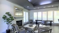 This single-floor space showcases a thoughtfully designed living area that blends contemporary elegance with functional comfort. The room's most striking architectural feature is its tray ceiling, finished in a deep charcoal grey that creates a dramatic contrast against the light grey walls, adding visual depth and a sense of elevated sophistication.

A classic fireplace with a marble-tiled surround and a white mantel serves as the room's focal point, complemented by a large, ornate mirror mounted above it that amplifies the sense of space and reflects natural light throughout the room. Recessed lighting in the tray ceiling provides warm, adjustable illumination.

Multiple windows dressed with white horizontal blinds line the walls, allowing generous amounts of natural light to filter in while offering views of the surrounding greenery. The seating arrangement features grey upholstered chairs with metallic chrome frames surrounding a substantial stone-topped coffee table, all anchored by a bold geometric black-and-white area rug. A sleek console table positioned near the windows adds additional functionality. The overall ambiance is polished, modern, and inviting.