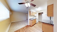 This single-floor space offers a functional and comfortable layout with a seamless flow between the kitchen and living area. The kitchen is equipped with warm honey-toned wood cabinetry, dark granite-style countertops, a stainless sink, white appliances including a refrigerator, electric range, and built-in microwave, all set atop hardwood-style flooring that adds a clean, classic aesthetic. A peninsula counter provides a natural divider between the kitchen and the adjacent living space, enhancing the open-concept feel. The living area transitions to plush carpeting, creating a cozy and inviting atmosphere. A ceiling fan with a light fixture is centrally positioned, providing both ambient lighting and air circulation. Neutral wall tones throughout keep the space feeling bright and versatile. A doorway visible from the main area leads to additional rooms, suggesting a practical and well-organized floor plan. Baseboard heating is present, ensuring warmth during cooler months. Overall, the space strikes a balance between functionality and comfort, making it well-suited for everyday living.