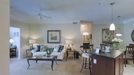 This single-floor residence is designed with an open-concept layout that promotes a sense of spaciousness and easy flow between living areas. The living room serves as the heart of the space, featuring light neutral walls, crown molding, and a ceiling fan that enhances comfort. Large framed artwork adorns the walls, adding a refined touch to the room's decor. The living area is anchored by plush seating and a classic wood coffee table, creating an inviting and well-appointed gathering space. Adjacent to the living room, a breakfast bar with dark countertops and bar seating provides a casual dining option, separating the living area from what appears to be a kitchen space. Light-toned tile flooring runs throughout, contributing to the bright and airy ambiance of the home. Warm, layered lighting from table lamps and overhead fixtures ensures the space feels welcoming at any time of day. A hallway visible from the living area leads to additional rooms, including what appears to be a bathroom with its own lighting and vanity. The overall design aesthetic is cohesive and elegant, combining dark wood accents with soft, neutral palettes to create a comfortable and stylish living environment.