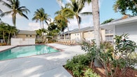 This single-level outdoor amenity space exudes a relaxed, resort-inspired atmosphere characteristic of Florida living. At its centerpiece is a generously sized swimming pool with crystal-clear water, bordered by a spacious concrete sun deck lined with rows of lounge chairs — ideal for soaking up the abundant natural sunlight. The pool area is enclosed by a white metal fence, providing both security and an open, airy feel.

Tall, swaying palm trees are planted throughout the grounds, creating natural shade and lending the space a distinctly tropical character. Thoughtfully designed landscaping beds filled with lush subtropical shrubs, flowering plants, and ornamental foliage frame the walkways and pool perimeter, adding layers of greenery and visual interest.

A prominent pergola structure with rich, warm-toned wood beams serves as a covered entrance, anchoring a transition zone between the outdoor grounds and interior facilities. Double glass-panel doors beneath the pergola provide access to interior spaces, while the open lattice roof of the pergola allows dappled light to filter through.

The surrounding buildings feature light yellow-beige horizontal siding and pitched gray rooflines, contributing to a cohesive coastal aesthetic. The overall ambiance is serene and inviting, combining well-maintained grounds with ample open-air space — well-suited for relaxation and outdoor recreation.