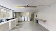 Located at 560 Beachway Ave in Keansburg, New Jersey, this single-floor shared laundry facility is designed to accommodate multiple users efficiently. The space is organized into two interconnected areas, each serving a distinct function within the laundry operation.

The main washing area houses a row of top-loading washing machines equipped with digital controls, positioned along one wall and accessible from an open central floor area finished with speckled terrazzo-style flooring. A dedicated section contains a matching row of front-vented dryers lined beneath a multi-pane window that allows natural light to enter and provides a view of the surrounding greenery. Fluorescent ceiling fixtures supplement the natural light throughout.

A secondary alcove area includes a utility sink for hand-washing or pre-treatment tasks, along with visible ductwork and utility connections. A wall-mounted folding table provides a convenient surface for sorting and folding laundry, situated near a waste bin for easy disposal. A fire extinguisher is mounted on the wall, and a solid metal door provides direct access to the exterior.

The facility maintains a clean, utilitarian aesthetic with white-painted walls and a straightforward layout that prioritizes functionality. Surveillance equipment is also present, contributing to the safety and security of the space.