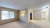 This single-floor residence offers a practical and well-organized layout designed for comfortable everyday living. The spacious living area is finished with plush wall-to-wall carpeting and features a textured ceiling, neutral warm-toned walls, and large windows fitted with horizontal blinds that allow ample natural light to fill the room. A wide picture window provides views of the outdoor surroundings, enhancing the bright and airy atmosphere.

A pass-through opening connects the living area directly to the kitchen, creating an open, communicative flow between the two spaces. The kitchen is equipped with warm oak cabinetry, speckled laminate countertops, a stainless steel sink positioned beneath a window, and black appliances including a gas range, built-in microwave, dishwasher, and refrigerator. The kitchen floor features light hardwood-style flooring, adding warmth to the space.

The bedroom is generously sized with soft carpeting, a ceiling fan with light fixture, a window for natural ventilation and light, and a closet for storage. Two doors visible from the entry area suggest additional closet or utility access.

The bathroom is thoughtfully appointed with wood-look flooring, a vanity featuring speckled countertops and an integrated sink, a large mirror, and a full tub-shower combination. The warm yellow-toned walls carry throughout the residence, creating a cohesive and inviting aesthetic.
