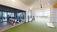This single-floor fitness and recreation facility is thoughtfully designed to support a wide range of wellness and social activities. The centerpiece of the space is a generously sized artificial turf area, ideal for functional training, recreational games such as cornhole, and group fitness activities. String lights suspended from the vaulted ceiling lend a warm, inviting ambiance to this open zone, which is framed by exposed wood beams and crisp white shiplap walls.

Adjacent to the turf, a dedicated spin and cardio studio is enclosed by floor-to-ceiling glass partition doors, housing a row of stationary cycling bikes alongside stability balls and additional cardio equipment. The glass enclosure allows natural light to flow between spaces while maintaining acoustic separation. A tiered dumbbell rack and medicine ball storage further equip the functional training area for strength-focused workouts.

The space transitions seamlessly into a semi-open lounge and dining area featuring herringbone brick flooring, high-top tables and chairs, pendant lighting, and a wall-mounted television — suited for casual gatherings or post-workout relaxation. A covered outdoor pavilion with slatted white screens, wooden dining tables, ceiling fans, and open views of the surrounding grounds completes the facility, blurring the boundary between indoor comfort and outdoor living.