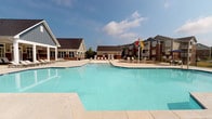 Located at 201 Babcock Lane in Duncan, South Carolina, this single-story outdoor amenity space centers around a generously sized swimming pool with clear, blue water and a smooth concrete surround. The pool features a gradually sloping entry point and a designated shallow water area, making it accessible for a range of swimmers. Depth markers are clearly posted along the pool's edge, and safety signage is prominently displayed throughout the enclosure.

The expansive sun deck provides ample space for lounging, with rows of poolside chaise lounges positioned to take full advantage of the open sky and natural sunlight. A splash pad area with colorful interactive water features adds a playful element to the space. Shaded seating areas, including Adirondack chairs and umbrella-covered tables, offer comfortable retreats from the sun.

The pool area is enclosed by a decorative black iron fence with ornamental finials, providing both security and a polished aesthetic. A well-maintained clubhouse structure with board-and-batten siding, gabled rooflines, and large windows lines one side of the pool, creating a welcoming backdrop. The surrounding landscaping includes neatly trimmed lawns and stone-accented garden beds, contributing to the overall well-kept and inviting atmosphere of the space.