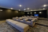 This screening room is designed to deliver a true cinema-quality experience in a polished, amenity-style setting. The main viewing area is anchored by a large wall-mounted display screen and a ceiling-mounted projector, offering multiple viewing options for streaming and entertainment. Rows of generously sized, neutral-toned lounge chairs and sectional seating are arranged in a tiered layout across patterned carpet flooring, ensuring clear sightlines from every seat. Accent pillows with bold geometric patterns add visual interest to the otherwise understated palette. The walls are finished in deep charcoal tones, enhancing the immersive atmosphere and minimizing light reflection during screenings. Recessed ceiling lighting and subtle wall-mounted sconces provide adjustable ambient illumination throughout the space. A side wall features additional display panels, while the room transitions seamlessly into a dedicated kitchenette area. The kitchenette is outfitted with sleek black cabinetry with gold hardware, dark countertops, a sink, microwave, and under-counter refrigerator, all set against a striking textured white accent wall. The overall design balances cinematic functionality with upscale comfort.