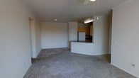 This single-floor space showcases an open-concept layout that integrates the living area and kitchen into one cohesive environment. The living area is finished with wall-to-wall carpeting in a neutral tone, creating a soft and understated foundation throughout the space. A ceiling fan is mounted overhead, contributing to both comfort and airflow.

The kitchen is defined by a wraparound breakfast bar with a dark countertop, which serves as a natural partition between the cooking and living zones while maintaining visual openness. Warm-toned wood cabinetry lines the upper kitchen walls, providing ample storage, and a white refrigerator anchors one end of the kitchen. Pendant lights hang above the bar area, adding focused illumination and a subtle design detail. Recessed lighting supplements the overhead fixtures, ensuring the space is well-lit throughout.

The walls are finished in a clean, neutral white, lending the space a bright and airy feel. A interior door visible along the back wall suggests access to additional areas of the unit. Overall, the layout is practical and efficient, with a straightforward design that prioritizes functionality and ease of movement.
