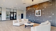 Located at 1179 E Main St in Columbus, Ohio, this single-floor commercial lobby space offers a generously proportioned, open-plan layout that makes a strong first impression. The interior is defined by light-toned hardwood flooring that runs throughout, lending warmth and continuity to the space.

A central focal point is a dramatic, textured feature wall finished in deep charcoal gray with an abstract geometric pattern, accented by warm bronze tones above. A statement circular pendant light anchors this area, casting a warm glow over a curated lounge arrangement of curved cream sofas and a round white coffee table. Recessed ceiling lighting supplements the ambient illumination throughout.

Adjacent to the main lounge is a secondary seating area furnished with sleek, tufted navy chairs on polished chrome frames beneath another circular pendant. This zone is bordered by floor-to-ceiling glass partitions with bold black steel framing and horizontal slatted screens, which allow natural light to filter in while defining distinct zones within the open space.

The entry corridor features clean white walls adorned with contemporary artwork and wall sconces, leading to large black-framed glass entrance doors that invite an abundance of natural light. Additional areas near the elevator core offer flexible seating with accent chairs in rich terracotta tones. The overall ambiance is modern, polished, and welcoming.