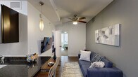 This efficiently designed one-bedroom, one-bathroom unit maximizes its compact footprint through a thoughtful, linear layout. The open living and dining area features hardwood-style flooring, a ceiling fan with integrated lighting, and a pendant light fixture, creating a warm and functional gathering space. A wall-mounted television and a built-in countertop area offer practical utility without sacrificing style.

The galley-style kitchen is well-appointed with dark espresso cabinetry, dark granite countertops, stainless steel appliances including a refrigerator, gas range, and microwave, and a stainless steel sink. The cabinetry extends to the ceiling, maximizing vertical storage capacity.

The bedroom accommodates a comfortable sleeping and working area, featuring carpeted flooring, a ceiling fan, and two closets—one standard and one additional storage closet—providing ample organization space. A window allows natural light to filter into the room.

The bathroom is finished with dark espresso cabinetry, granite countertops, a wall-mounted medicine cabinet, large-format dark tile flooring, and a full tub-shower combination with subway tile surround. Throughout the unit, a consistent neutral color palette and modern finishes contribute to a cohesive, contemporary aesthetic.