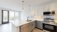 This well-designed one-bedroom, one-bathroom unit offers a thoughtful layout across a single floor, making efficient use of every square foot. The open-concept kitchen and living area serves as the heart of the space, where contemporary two-tone cabinetry — white upper cabinets paired with sage-gray lowers — is complemented by white quartz countertops, a textured tile backsplash, and a full suite of stainless steel appliances including a range, microwave, dishwasher, and refrigerator. Gold-toned pendant lights hang above the kitchen peninsula, adding a warm accent to the modern aesthetic. Light wood-tone flooring flows throughout the main living areas, and large sliding glass doors lead to a private balcony, filling the living room with natural light and outdoor views. A built-in desk nook with upper cabinetry provides a dedicated workspace just off the kitchen. The bedroom features plush carpet, a natural light-filled window, and a stylish gray panel door. It connects directly to the bathroom and a well-organized walk-in closet equipped with multi-tiered shelving and hanging rods. The bathroom offers a sleek vanity with dark hardware, a large-format tile walk-in shower with frameless glass enclosure, and penny tile flooring, completing this polished and functional home.