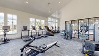 This expansive fitness center is designed to accommodate a wide range of workout styles within a single, open-plan space. The room's most striking architectural feature is its dramatically vaulted ceiling, which rises to a considerable height and contributes to an airy, open atmosphere throughout. Crisp white walls and large multi-pane windows line the perimeter, flooding the interior with natural light and offering views of an outdoor pool area and surrounding palm trees.

The flooring throughout is covered in patterned blue-grey carpet tile, providing a comfortable and functional surface underfoot. The fitness center is divided into distinct workout zones. A dedicated cardio area houses a row of Matrix treadmills and stationary bikes positioned directly in front of the large windows, alongside elliptical trainers and a rowing machine. A strength training zone features a cable machine system and selectorized equipment with blue upholstery. A functional training area includes a well-stocked rack with kettlebells, resistance bands, battle ropes, medicine balls, and a plyo box, as well as a squat rack with a bench. A wall-mounted flat-screen TV and a separate glass-walled cycling studio accessible via black-framed doors round out the facility.