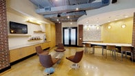 This single-floor space showcases a compelling blend of industrial and contemporary design elements. The open layout is anchored by polished concrete floors that reflect warm ambient light throughout the room, contributing to an inviting atmosphere. Exposed brick columns add textural contrast and architectural character, framing a set of sleek black double doors with frosted glass panels that serve as a striking focal point.

The ceiling features an exposed industrial framework with track lighting, adding a raw, urban aesthetic while providing flexible, directed illumination. A dedicated lounge area is furnished with modern swivel chairs in warm brown tones, arranged around a low-profile rectangular coffee table, creating an ideal setting for casual gatherings or meetings.

Adjacent to the lounge, a dining or conference area offers counter-height seating at a long white table, illuminated by pendant lighting with amber-toned globes. A mosaic tile accent wall in the background adds depth and visual interest to this zone.

Built-in cabinetry and open shelving along one wall provide functional storage, decorated with metallic spherical accents and a contemporary vase. Together, these elements create a cohesive, well-appointed space suited for both social and professional use.