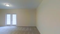This single-floor space presents a clean, open layout finished in warm neutral tones throughout. The living area is generously proportioned, offering a sense of openness enhanced by plush wall-to-wall carpeting in a light beige hue that complements the soft cream-colored walls. Smooth ceilings with flush-mount lighting provide even illumination, while white painted baseboards add a crisp, finished detail to each room.

A standout feature of the main living area is the set of French doors, framed in white and fitted with frosted glass panels, which allow natural light to filter into the space while providing access to the exterior. An additional window along one wall further brightens the interior, contributing to the airy, welcoming atmosphere.

The layout connects fluidly between rooms, with a hallway visible from the main living area suggesting additional rooms beyond. The space is entirely vacant, making it easy to envision a variety of furniture arrangements and interior configurations. Overall, the space offers a straightforward, functional design with neutral finishes that provide a versatile backdrop suitable for a wide range of personal styles and uses.