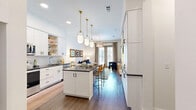 Located at 1010 Singleton Blvd in Dallas, TX, this one-bedroom, one-bathroom unit offers a thoughtfully designed layout that maximizes every square foot. The open-concept kitchen and living area creates a seamless flow, anchored by a central kitchen island with dark countertops and bar seating. The kitchen is outfitted with white shaker cabinetry, a subway tile backsplash, stainless steel appliances, and gold-accented pendant lighting that adds a contemporary touch. A built-in nook near the refrigerator provides additional storage and organization. The living room benefits from large floor-to-ceiling windows and a glass door that invite an abundance of natural light and offer views of the surrounding Dallas cityscape. A stylish chandelier serves as a focal point overhead. Hardwood-style flooring runs throughout the main living areas, lending warmth and continuity. The bedroom is a comfortable retreat, featuring a large window, ample natural light, and direct access to a generously sized walk-in closet with built-in shelving and hanging rods. The bathroom is finished with gray cabinetry, dark countertops, large-format tile flooring, and a well-lit vanity mirror. A hallway connects the private and shared spaces efficiently, completing this well-appointed urban residence.