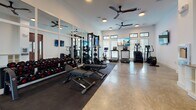 Located at 2500 Gault Way Dr in Summerville, SC, this single-floor fitness center is thoughtfully designed to support a wide range of workout routines. The facility is divided into two distinct training areas, each tailored to different exercise disciplines.

The strength training zone features a full power rack with barbell attachments, a comprehensive dumbbell rack spanning multiple weight increments, adjustable benches, and floor-mounted weight plates — all set atop protective rubber matting. Full-length framed mirrors line the walls, allowing for proper form checks, while wall-mounted flat-screen TVs provide entertainment during workouts. Ceiling fans maintain air circulation throughout.

The functional training area offers a more open layout with cushioned interlocking floor tiles, suspended TRX straps for bodyweight training, a rowing machine, and additional cardio equipment including treadmills, an elliptical, and a stationary bike. Large windows with shutters flood this space with natural light and offer views of the surrounding landscape. Elevated transom windows add further brightness and visual openness.

The entry corridor connecting both zones features polished concrete floors, warm wood-framed glass doors, and built-in white cabinetry, lending a clean and organized transition between spaces. The overall design balances functionality with a modern, welcoming aesthetic.