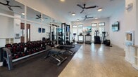 Located at 2500 Gault Way Dr in Summerville, SC, this single-floor fitness center is thoughtfully designed to support a wide range of workout routines. The facility is divided into two distinct training areas, each tailored to different exercise disciplines.

The strength training zone features a full power rack with barbell attachments, a comprehensive dumbbell rack spanning multiple weight increments, adjustable benches, and floor-mounted weight plates — all set atop protective rubber matting. Full-length framed mirrors line the walls, allowing for proper form checks, while wall-mounted flat-screen TVs provide entertainment during workouts. Ceiling fans maintain air circulation throughout.

The functional training area offers a more open layout with cushioned interlocking floor tiles, suspended TRX straps for bodyweight training, a rowing machine, and additional cardio equipment including treadmills, an elliptical, and a stationary bike. Large windows with shutters flood this space with natural light and offer views of the surrounding landscape. Elevated transom windows add further brightness and visual openness.

The entry corridor connecting both zones features polished concrete floors, warm wood-framed glass doors, and built-in white cabinetry, lending a clean and organized transition between spaces. The overall design balances functionality with a modern, welcoming aesthetic.