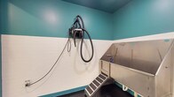 This self-service dog wash facility is a purpose-built grooming space designed with both functionality and ease of use in mind. The room features a two-tone color scheme, with crisp white tile wainscoting along the lower half of the walls and a vibrant teal paint above, creating a clean and cheerful environment. The centerpiece of the space is a deep stainless steel washing tub, elevated for comfortable use and equipped with a retractable coiled sprayer hose, allowing for thorough and efficient bathing. A set of non-slip entry steps provides safe and easy access for dogs of all sizes. Mounted on the adjacent wall is a professional-grade forced-air dryer with a flexible hose and multiple attachments, ensuring pets can be thoroughly dried after their bath. The dark, sealed flooring is designed for easy cleaning and drainage, complementing the room's overall hygienic design. The layout is compact yet efficiently organized, with all essential grooming tools within arm's reach. This space provides everything needed for a complete pet grooming session in a well-maintained, user-friendly environment.