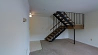 This two-floor space makes a strong first impression with its open, modern staircase, which serves as a central architectural feature on the main level. The staircase is constructed with carpeted treads and sleek black metal railings and vertical balusters, creating a visually open and industrial aesthetic that contrasts cleanly against the white walls. A wooden handrail adds a warm touch to the otherwise bold metalwork. The main level is finished with wall-to-wall carpet in a neutral beige tone, contributing to a soft and comfortable underfoot feel throughout the space. The white walls are clean and minimalist, offering a versatile backdrop that suits a variety of interior styles. A ceiling-mounted light fixture provides overhead illumination on the main floor, keeping the space adequately lit. The open-riser staircase design allows light and sightlines to pass through, enhancing the sense of openness between the two floors. The upper level is visible from below, with the staircase landing offering a glimpse of the continued layout above. Overall, the space presents a functional and straightforward design with a clean, modern character.
