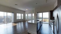 This single-floor space showcases a thoughtfully designed open-concept layout that seamlessly connects the kitchen and living areas. Dark hardwood flooring runs throughout, providing a rich, warm contrast to the bright white walls and ceilings. The kitchen is anchored by a large island with a crisp white countertop and a built-in sink, complemented by dark cabinetry that adds a contemporary touch. Pendant lighting above the island enhances the modern aesthetic, while recessed and flush-mounted ceiling fixtures provide ambient illumination throughout.

The living area is generously proportioned, benefiting from abundant natural light streaming through multiple windows fitted with horizontal blinds. A sliding glass door opens to a balcony, offering elevated outdoor views of the surrounding landscape and a distant cityscape, making it a notable feature of the space. The open floor plan creates a fluid, airy atmosphere that encourages easy movement between living and cooking areas. Overall, the space presents a clean, modern aesthetic defined by its neutral palette, quality finishes, and strong connection to natural light and outdoor scenery.