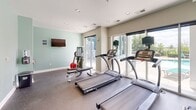 This fitness center is a thoughtfully designed exercise space finished with neutral beige and soft sage green walls, recessed lighting, and durable rubber-flooring throughout. Large floor-to-ceiling windows and glass sliding doors line one wall, flooding the room with natural light and offering direct views of an outdoor pool and surrounding greenery — creating an energizing atmosphere for workouts.

The space is outfitted with a comprehensive selection of cardio equipment, including multiple SportsArt treadmills, an elliptical trainer, a stationary cycle, and a rowing machine. A wall-mounted flat-screen television provides entertainment during workouts. Strength and functional training needs are served by a multi-station cable machine, a workout bench, and a medicine ball, making the facility well-rounded for a variety of fitness routines.

Structural columns with decorative green accents add an architectural touch, while a mirrored glass partition wall and a glass-paneled entry door contribute to an open, airy feel. Ventilation is provided through ceiling-mounted HVAC vents, and a clearly marked emergency exit ensures safety. The overall design balances functionality with a clean, modern aesthetic, offering users a comfortable and practical environment to support their fitness goals.