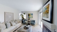 Located at 15639 Avenida Alcocofa in San Diego, this single-story home offers a thoughtfully designed interior that balances comfort and style. The living room serves as the centerpiece of the space, showcasing a clean, neutral color palette with soft white and warm greige tones across the walls and ceiling. Plush, light-colored carpeting underfoot adds warmth and texture to the room.

Natural light fills the space generously, thanks to multiple windows fitted with white plantation shutters and a large sliding glass door that opens to an outdoor view, bringing the California sunshine indoors. The sliding door also provides a seamless visual connection to the exterior greenery beyond.

The living room is anchored by a comfortable white sofa and complemented by a pair of soft blue accent chairs arranged around a round, decorative coffee table. A patterned area rug grounds the seating arrangement, while carefully placed indoor plants and curated wall art add color and personality to the space. A fireplace is partially visible along one wall, contributing an additional element of comfort and charm. The overall ambiance is bright, airy, and inviting.