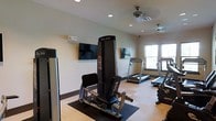 This single-floor fitness room offers a functional and comfortable workout environment designed to support a range of exercise routines. The space features a combination of strength training and cardiovascular equipment, including a Body-Solid cable/leg press machine, multiple treadmills, and additional cardio equipment positioned near a large window that allows natural light to fill the room. The light-toned flooring, accented with darker rubber mat sections beneath the equipment, provides both a clean aesthetic and practical safety underfoot. Two wall-mounted flat-screen televisions are positioned for easy viewing during workouts, enhancing the overall exercise experience. Ceiling fans help maintain comfortable airflow throughout the space. The neutral-toned walls and recessed lighting contribute to a bright, open atmosphere. The layout is thoughtfully arranged to allow adequate space between machines, ensuring ease of movement and safe use of equipment. Overall, the fitness room presents a practical and well-maintained facility suited for daily exercise needs.