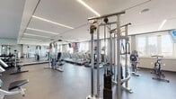 Located at 6804 Tides Rd in New York, NY, this well-equipped fitness center offers a comprehensive workout environment designed to support a wide range of exercise routines. The space is housed on a single floor and features a clean, modern aesthetic with neutral gray walls, a dark rubberized flooring surface, and a suspended ceiling fitted with sleek linear LED lighting that ensures consistent, bright illumination throughout.

The cardio zone is prominently featured and includes multiple treadmills, elliptical trainers, and a stationary bike, all positioned along windowed walls that allow natural light to filter in and provide views of the surrounding exterior. Wall-mounted flat-screen televisions are conveniently placed for use during cardio sessions. Full-length mirrors line portions of the walls, visually expanding the space and allowing users to monitor their form.

The strength training area is equally well-appointed, offering cable machine stations with adjustable pulley systems, weight benches, and a functional trainer suitable for a variety of resistance exercises. A dedicated free weights section includes a tiered kettlebell rack stocked with multiple weight increments, complemented by open floor space for functional training. Built-in shelving provides organized storage along one wall. The overall layout is spacious and thoughtfully arranged, promoting an efficient and comfortable workout experience.