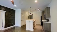 This single-floor space showcases a thoughtfully designed open-concept layout that seamlessly blends functionality with modern aesthetics. The kitchen serves as the centerpiece, featuring a sleek white island with a built-in sink and clean shaker-style paneling, complemented by light quartz countertops. Striking pendant lights with deep red accents hang above the island, adding a bold design touch against the bright white ceiling. Track lighting and recessed ceiling lights ensure the space is well-illuminated throughout. Stainless steel appliances, including a refrigerator and dishwasher, lend a polished, contemporary feel to the kitchen. The flooring throughout is polished concrete, offering a sophisticated industrial character that contrasts beautifully with the crisp white walls and cabinetry. A textured dark accent wall, reminiscent of weathered wood or slate, adds warmth and visual depth to one side of the room. A louvered closet door and a smart thermostat panel are neatly integrated into the wall, pointing to practical, well-considered design choices. The open layout allows for a generous flow between the kitchen and living areas, creating an inviting and versatile living environment ideal for both everyday living and entertaining.