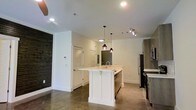 This single-floor space showcases a thoughtfully designed open-concept layout that seamlessly blends functionality with modern aesthetics. The kitchen serves as the centerpiece, featuring a sleek white island with a built-in sink and clean shaker-style paneling, complemented by light quartz countertops. Striking pendant lights with deep red accents hang above the island, adding a bold design touch against the bright white ceiling. Track lighting and recessed ceiling lights ensure the space is well-illuminated throughout. Stainless steel appliances, including a refrigerator and dishwasher, lend a polished, contemporary feel to the kitchen. The flooring throughout is polished concrete, offering a sophisticated industrial character that contrasts beautifully with the crisp white walls and cabinetry. A textured dark accent wall, reminiscent of weathered wood or slate, adds warmth and visual depth to one side of the room. A louvered closet door and a smart thermostat panel are neatly integrated into the wall, pointing to practical, well-considered design choices. The open layout allows for a generous flow between the kitchen and living areas, creating an inviting and versatile living environment ideal for both everyday living and entertaining.