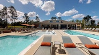 Located at 5090 Tradeway Dr in Leland, NC, this single-level amenity clubhouse serves as the social and recreational hub of a modern residential community. The centerpiece of the outdoor space is a generously sized swimming pool with crystal-clear water, complemented by a separate spa and a shallow sun-shelf area ideal for relaxing. The expansive pool deck is lined with chaise lounges and shade umbrellas, providing ample space for sunbathing and outdoor gatherings. A charming brick-constructed fire pit with seating adds a warm, inviting focal point to the deck area. The clubhouse building features a distinctive navy and wood-tone exterior with a standing-seam metal roof accent. A covered outdoor veranda with a warm wood-paneled ceiling, ceiling fans, and comfortable wicker seating with bold orange cushions offers a shaded retreat overlooking the pool. A ping-pong table on the covered patio adds a recreational element to the space. Beyond the pool area, a full tennis and pickleball court provides additional outdoor recreation options. The entire amenity complex is enclosed by decorative iron fencing, with tall pines framing the skyline and the broader residential buildings visible in the background, creating a cohesive and well-appointed community environment.