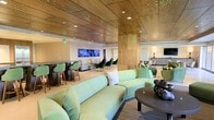 This contemporary lounge space is designed with a refined aesthetic that blends warm materials with modern flair. The standout architectural feature is the rich wood-paneled ceiling, which spans the entire area and is complemented by clusters of subtle green accent lighting that cast a distinctive, ambient glow throughout the room. The lighting design adds a dynamic and upscale character to the space.

The main seating area is anchored by a large, curved sage-green velvet sofa paired with a sleek, round sculptural coffee table, creating an inviting central gathering zone. Plush green accent chairs are thoughtfully distributed throughout, maintaining a cohesive color palette and ensuring comfortable seating across multiple areas.

A built-in bar or high-top counter runs along one side of the room, accompanied by matching green bar stools, making it an ideal spot for social interaction. Multiple large-screen displays are mounted along the wall behind the counter, adding a media-forward, entertainment-ready element to the space.

Warm column accents with decorative perforated detailing divide the space subtly while maintaining an open, flowing layout. The overall ambiance is polished and welcoming, combining contemporary design elements with comfortable furnishings to create a versatile space well-suited for lounging, socializing, or events.