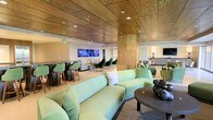 This contemporary lounge space is designed with a refined aesthetic that blends warm materials with modern flair. The standout architectural feature is the rich wood-paneled ceiling, which spans the entire area and is complemented by clusters of subtle green accent lighting that cast a distinctive, ambient glow throughout the room. The lighting design adds a dynamic and upscale character to the space.

The main seating area is anchored by a large, curved sage-green velvet sofa paired with a sleek, round sculptural coffee table, creating an inviting central gathering zone. Plush green accent chairs are thoughtfully distributed throughout, maintaining a cohesive color palette and ensuring comfortable seating across multiple areas.

A built-in bar or high-top counter runs along one side of the room, accompanied by matching green bar stools, making it an ideal spot for social interaction. Multiple large-screen displays are mounted along the wall behind the counter, adding a media-forward, entertainment-ready element to the space.

Warm column accents with decorative perforated detailing divide the space subtly while maintaining an open, flowing layout. The overall ambiance is polished and welcoming, combining contemporary design elements with comfortable furnishings to create a versatile space well-suited for lounging, socializing, or events.